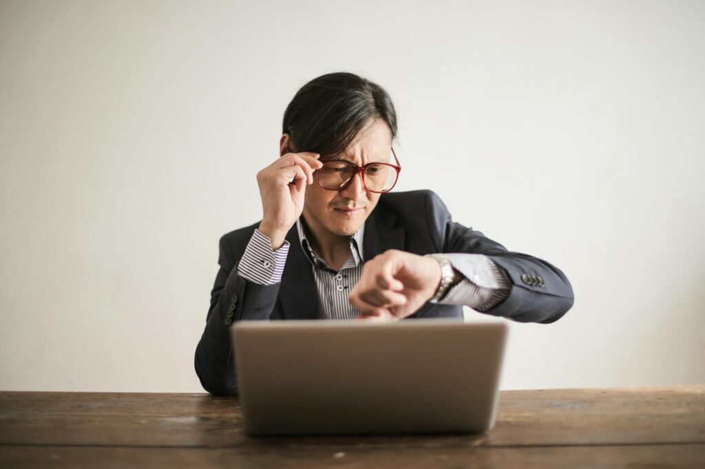 Young frowning man in suit and glasses looking at wristwatch while waiting for appointment sitting at desk with laptop